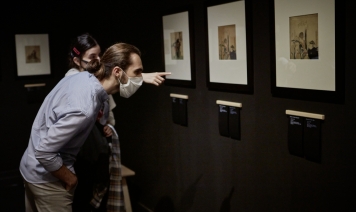  Dwójka uczestników projektu “Spektrum” zwiedza wystawę Hokusaia. Młoda kobieta wskazuje palcem na drzeworyt przedstawiający kobietę i dwóch mężczyzn, a stojący przed nią chłopak schyla się, by przeczytać podpis pod innym drzeworytem.