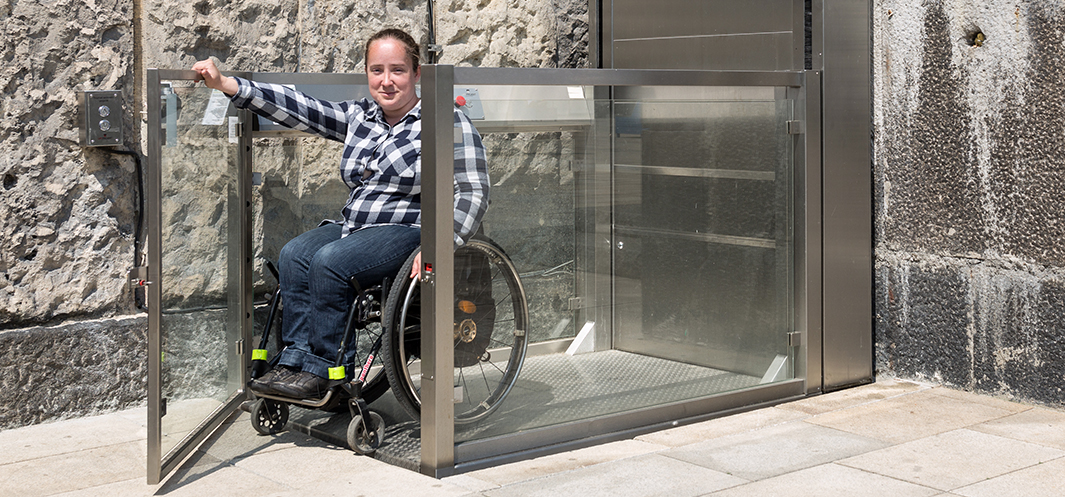 Uśmiechnięta młoda kobieta na wózku inwalidzkim korzysta z windy dla osób z niepełnosprawnościami znajdującej się na zewnątrz Gmachu Głównego muzeum. 