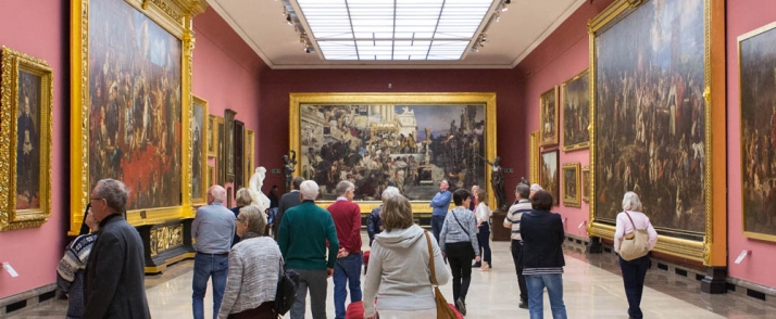 Wnętrze sali wystawowej w Galerii Sztuki Polskiej XIX wieku. Osoby zwiedzające muzeum przyglądają się wielko- i małoformatowym obrazom umieszczonym w sali.