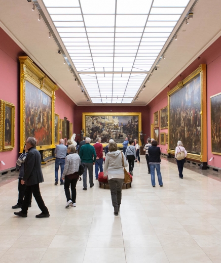 Wnętrze sali wystawowej w Galerii Sztuki Polskiej XIX wieku. Osoby zwiedzające muzeum przyglądają się wielko- i małoformatowym obrazom umieszczonym w sali.