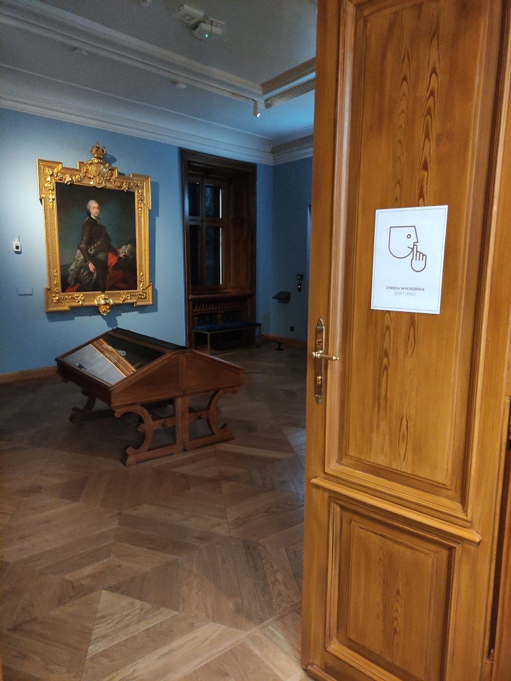 Sala w Muzeum Książąt Czartoryskich. Przed salą uchylone drzwi, na których przyklejona jest kartka z napisem “strefa wyciszenia” i piktogramem przedstawiającym człowieka wykonującym gest uciszania. 