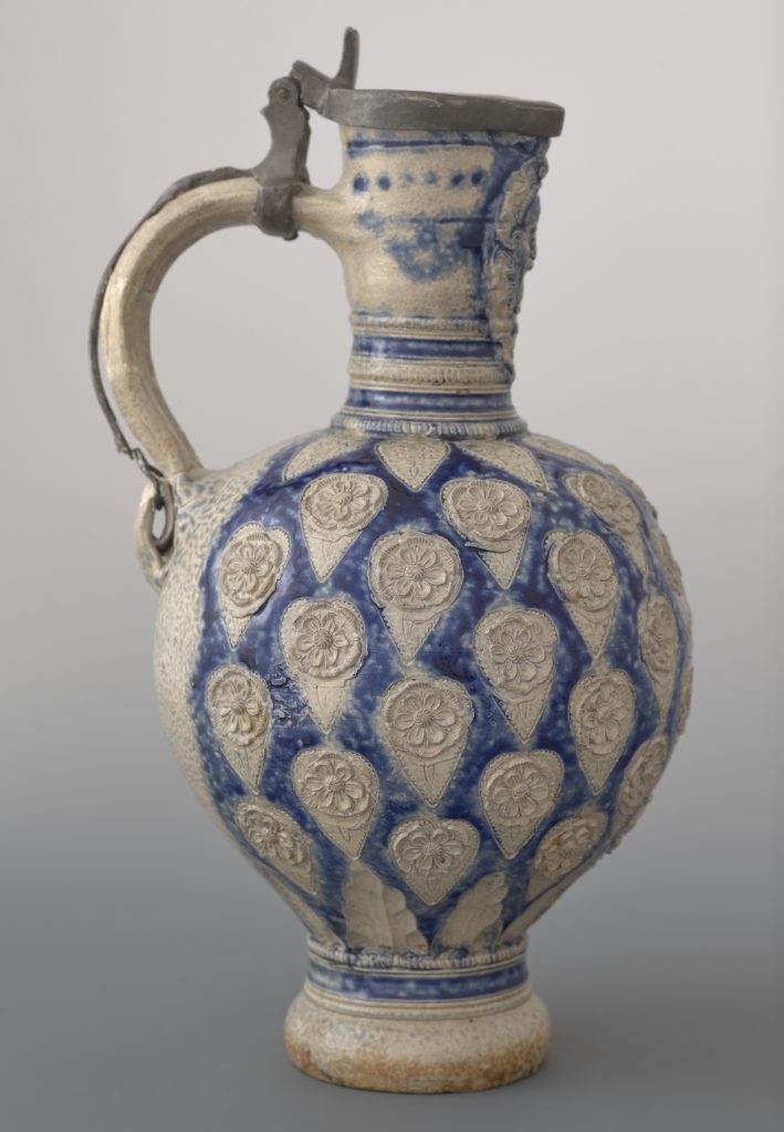 Duży ceramiczny dzban z uchwytem i metalową pokrywką, kremowo-beżowa glazura z intensywnie niebieskimi polami i powtarzającymi się reliefowymi kroplowymi medalionami z kwiatowymi motywami na korpusie; szyjka ozdobiona poziomymi paskami i detalami, metalowy zawias pokrywki przymocowany do uchwytu oraz widoczne ślady zużycia przy podstawie na neutralnym szarym tle.