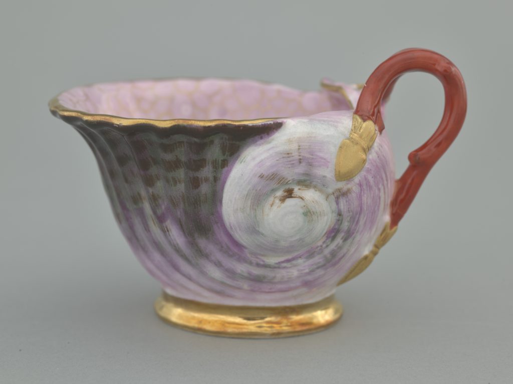 Zbliżenie fragmentu ozdobnego porcelanowego naczynia z czerwonym uchwytem po prawej, złotym rantem i podstawą, spiralnym fioletowo‑różowym malowaniem na korpusie oraz jasnoróżowym nakrapianym wnętrzem.