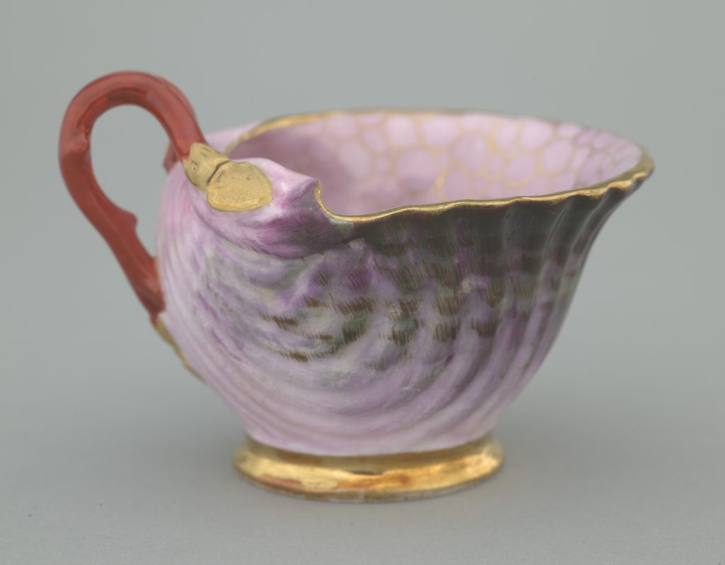 Zbliżenie na porcelanowy dzbanuszek o skorupkowym, falistym kształcie z różowo‑fioletowym nakrapianym szkliwem, złoconą krawędzią i podstawą oraz czerwonym zakrzywionym uchwytem na szarym tle.