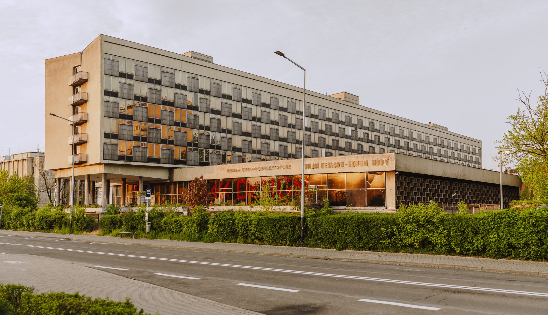 Duży betonowy modernistyczny budynek o wydłużonej, prostokątnej bryle z rzędami regularnych okien odbijających pomarańczowe światło, małymi balkonami po lewej stronie, przeszklonym parterem z widocznym napisem na fasadzie, przed nim żywopłot i krzewy, chodnik, pusta jezdnia oraz kilka latarni ulicznych.