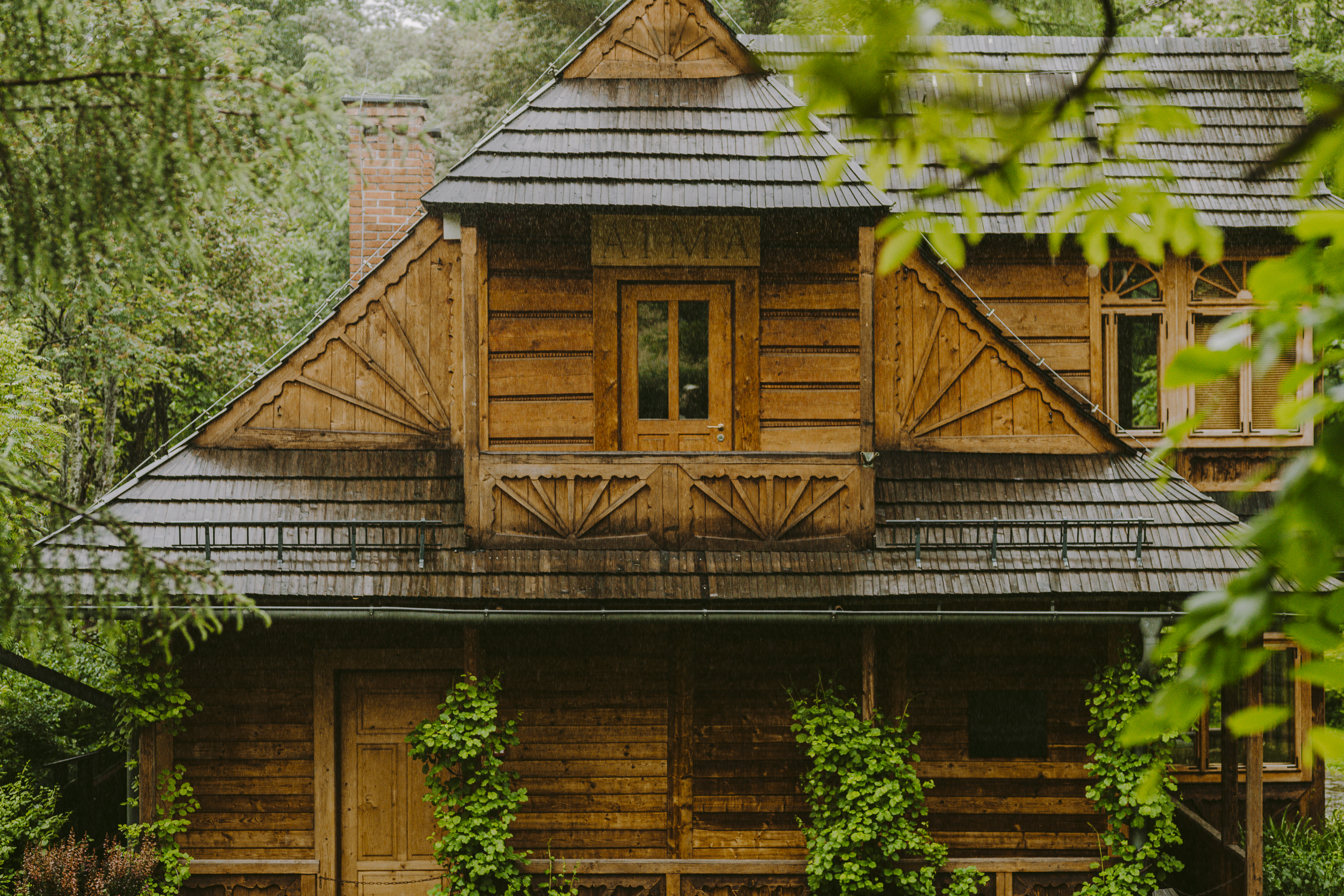 Drewniany domek w lesie z ciemnym gontowym dachem i ozdobnymi rzeźbieniami, centralnym drzwiowym oknem na poddaszu z małym balkonem, ceglanym kominem po lewej, dodatkowymi oknami po prawej oraz pnączami wspinającymi się po elewacji, uchwycony podczas padającego deszczu.