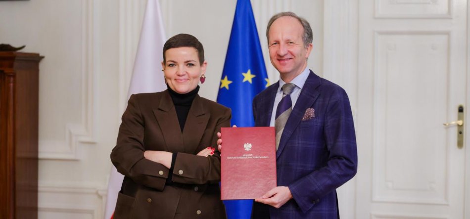 Dwie osoby w formalnych strojach stoją w jasnym pomieszczeniu przed flagami Polski i Unii Europejskiej; kobieta w brązowej marynarce z założonymi rękami, mężczyzna w kratowanej granatowej marynarce trzyma czerwony dokument z godłem Polski.