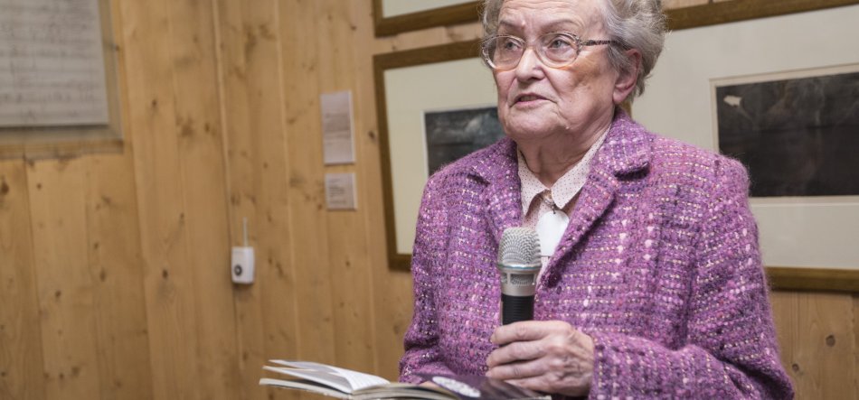 Starsza kobieta z krótkimi siwymi włosami i okularami, ubrana w fioletowo-różowy żakiet, trzyma mikrofon i otwartą książkę stojąc przed drewnianą ścianą z oprawionymi obrazami.