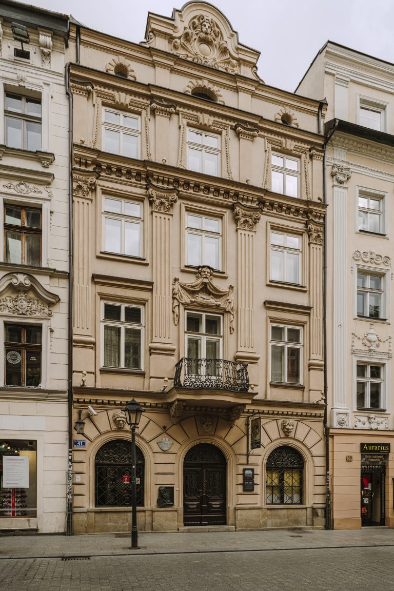 Widok fasady jasnobeżowej, ozdobnej kamienicy z kilkoma kondygnacjami okien nad parterowymi, łukowymi wejściami, rzeźbionymi pilastrami i stiukowymi gzymsami, żeliwną balkonową balustradą nad centralnym oknem, dekoracyjnymi maskami i medalionem w szczycie oraz sąsiadującymi kamienicami po bokach, na chodniku przed budynkiem latarnia i brukowana ulica.