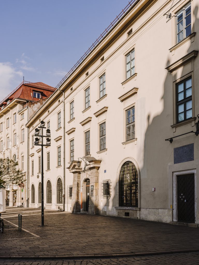 Jasna fasada wielopiętrowego budynku z regularnie rozmieszczonymi prostokątnymi oknami, kamiennym portalem wejściowym i łukowymi kratami na parterze, czerwonym dachem po lewej, słupem z reflektorami na brukowanym deptaku i dużym cieniem padającym na ścianę.