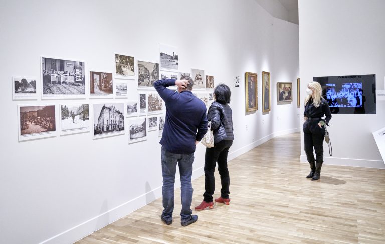 Wnętrze galerii z białymi ścianami i jasną drewnianą podłogą, na lewej ścianie uporządkowana wystawa starych czarno‑białych fotografii i kilka oprawionych obrazów, przy nich dwie osoby (mężczyzna w granatowym swetrze drapiący się po głowie i kobieta w czarnej kurtce z torebką i czerwonymi butami) oglądające prace, a po prawej stoi kobieta z blond włosami w czarnym ubraniu i białej maseczce obok ekranu wyświetlającego niebieski filmowy obraz.