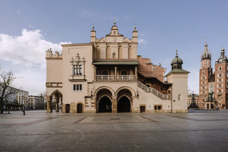 Rozległy brukowany plac przed dużym ozdobnym budynkiem o kremowej fasadzie z trzema arkadami na parterze, górnym balkonem z balustradą, zewnętrznymi schodami po prawej, kopułką i wieżyczkami, a po prawej w tle widoczne wysokie ceglaste wieże kościelne pod jasnym niebem z nielicznymi chmurami.