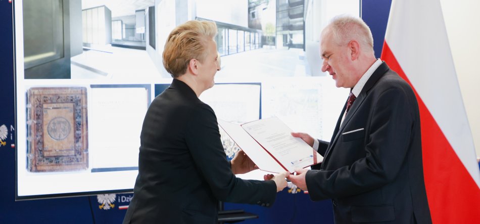 Osoba po lewej o krótkich jasnych włosach w czarnym garniturze podaje teczkę z dokumentem osobie po prawej o krótkich siwych włosach w ciemnym garniturze, w tle duży ekran z grafikami i widoczna flaga Polski.