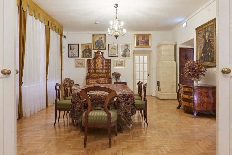 Elegancka, klasyczna jadalnia widoczna przez rozwarte białe drzwi z długim stołem przykrytym wzorzystym obrusem i drewnianymi krzesłami z zielonymi siedziskami, po lewej długie białe i musztardowe zasłony, u góry ozdobny żyrandol, na tylnej ścianie galerie portretów i obrazów nad rzeźbioną komodą, po prawej wysoki biały kaflowy piec i kolejna zdobna komoda z wazonem suszonych kwiatów, całość na jasnej parkietowej podłodze.