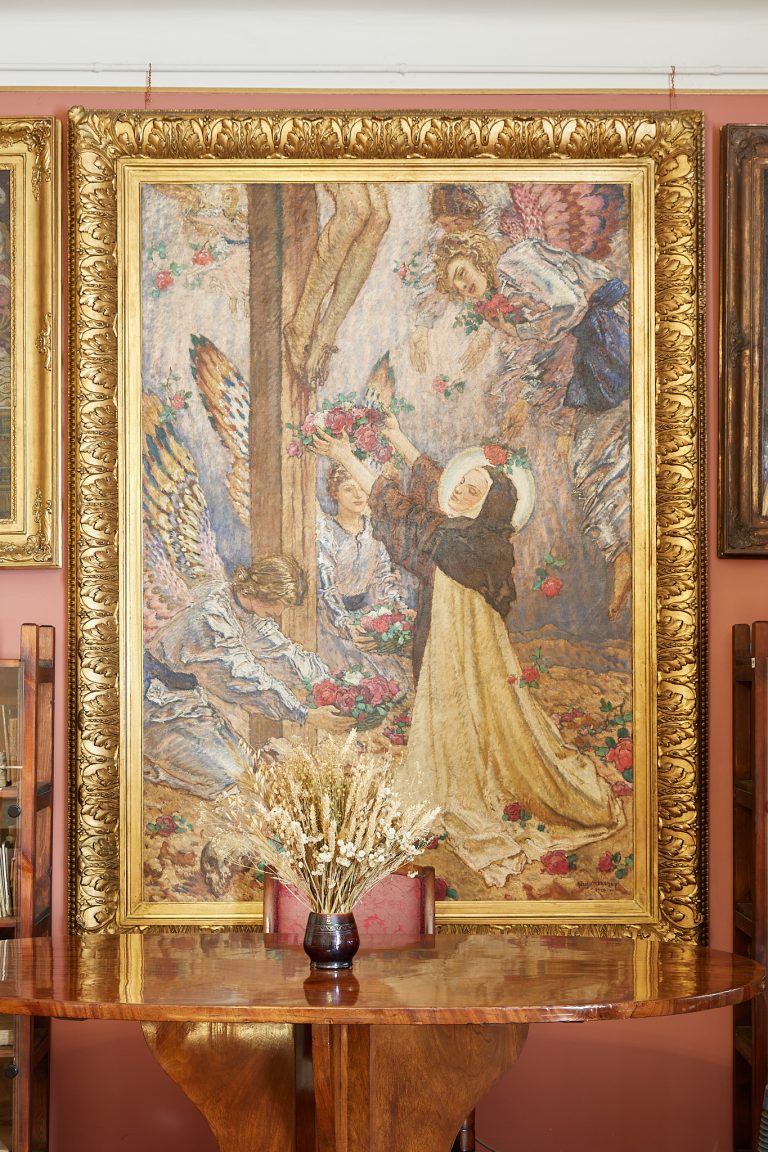 Pionowy widok dużego kolorowego obrazu w bogato rzeźbionej złotej ramie wiszącego na łososiowo-różowej ścianie, przedstawiającego postacie z aureolami i skrzydłami oraz inne osoby trzymające wieńce i kwiaty, a na pierwszym planie przed obrazem okrągły drewniany stół z ciemnym wazonem wypełnionym suszonymi trawami i kwiatami.