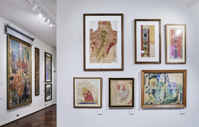 Biała ściana galerii z oprawionymi rysunkami i ilustracjami w drewnianych ramach — u góry centralnie duży pionowy arkusz z ornamentem w odcieniach czerwieni i złota, po prawej dwie wąskie pionowe prace z kolorowymi geometrycznymi wzorami, poniżej trzy mniejsze ramy: po lewej kwadrat z motywem roślinnym i czerwonym trójkątem, w środku okrągły motyw z czerwonymi elementami w złotej ramie, po prawej prostokątna praca z niebiesko‑zielonymi kwiatami na geometrycznym tle; po lewej kadr sali wystawy stałej