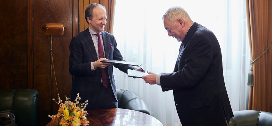 Dwaj mężczyźni w garniturach stoją naprzeciwko siebie w eleganckim gabinecie, wymieniając teczki i dokumenty — mężczyzna po lewej się uśmiecha, przed nimi okrągły drewniany stół z bukietem kwiatów, w tle zasłony, okno, zielone skórzane fotele i stojąca lampa.