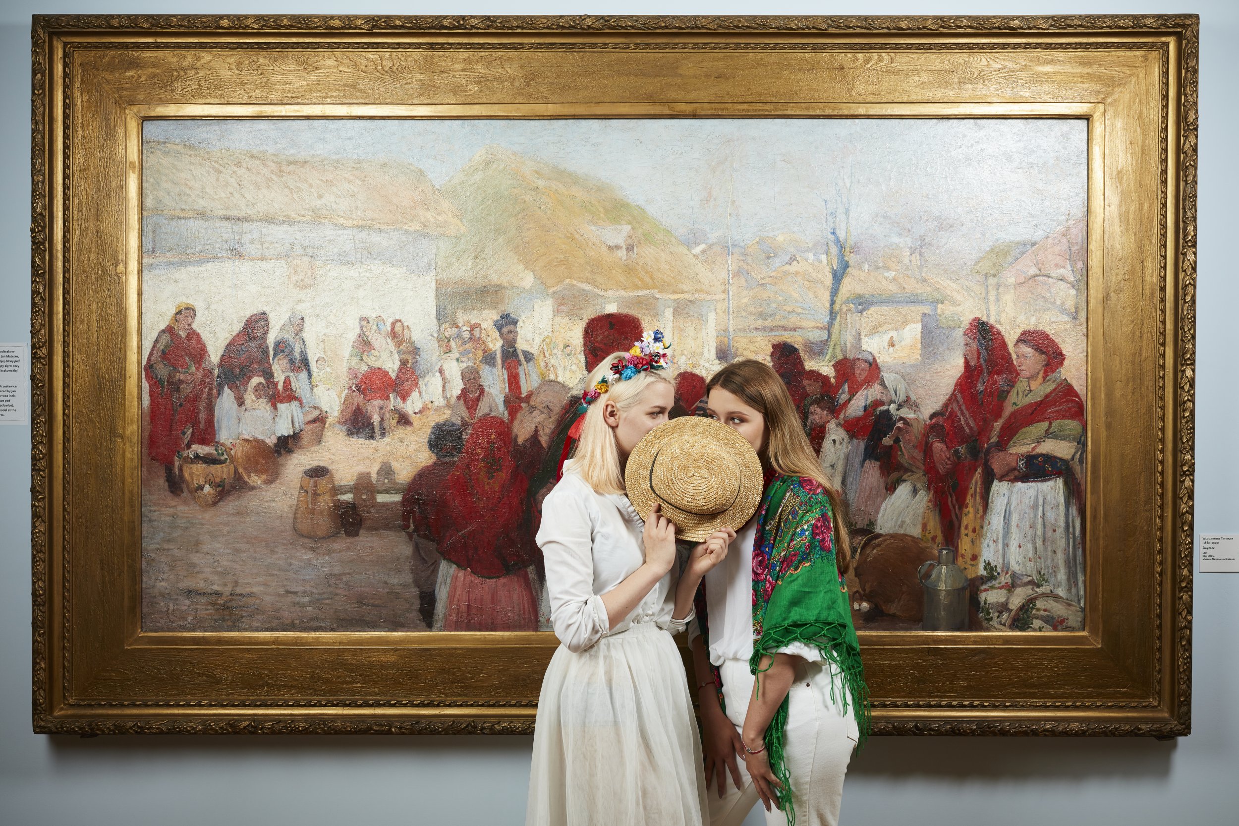 Dwie młode kobiety stoją przed dużym obrazem w ozdobnej złotej ramie na ścianie muzeum: jedna w białej sukience z wianuszkiem z kwiatów trzyma słomkowy kapelusz zasłaniający usta, druga w białych spodniach i zielonym, kwiatowym szalu pochyla się obok, a w malowidle widać wiejską scenę z wieloma postaciami w czerwonych chustach i chatami w tle.