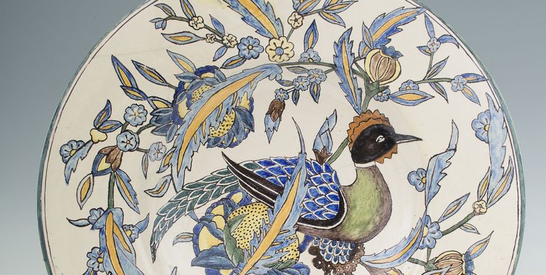 Okrągły ceramiczny talerz z centralnym malunkiem ptaka o czarnej głowie, zielonym tułowie i niebieskich łuskowatych skrzydłach, otoczony stylizowanymi pnączami i kwiatami w odcieniach niebieskiego, żółtego i zielonego na kremowym tle.