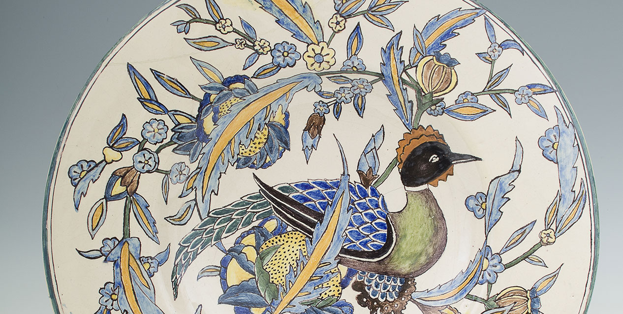 Okrągły ceramiczny talerz z centralnym malunkiem ptaka o czarnej głowie, zielonym tułowie i niebieskich łuskowatych skrzydłach, otoczony stylizowanymi pnączami i kwiatami w odcieniach niebieskiego, żółtego i zielonego na kremowym tle.