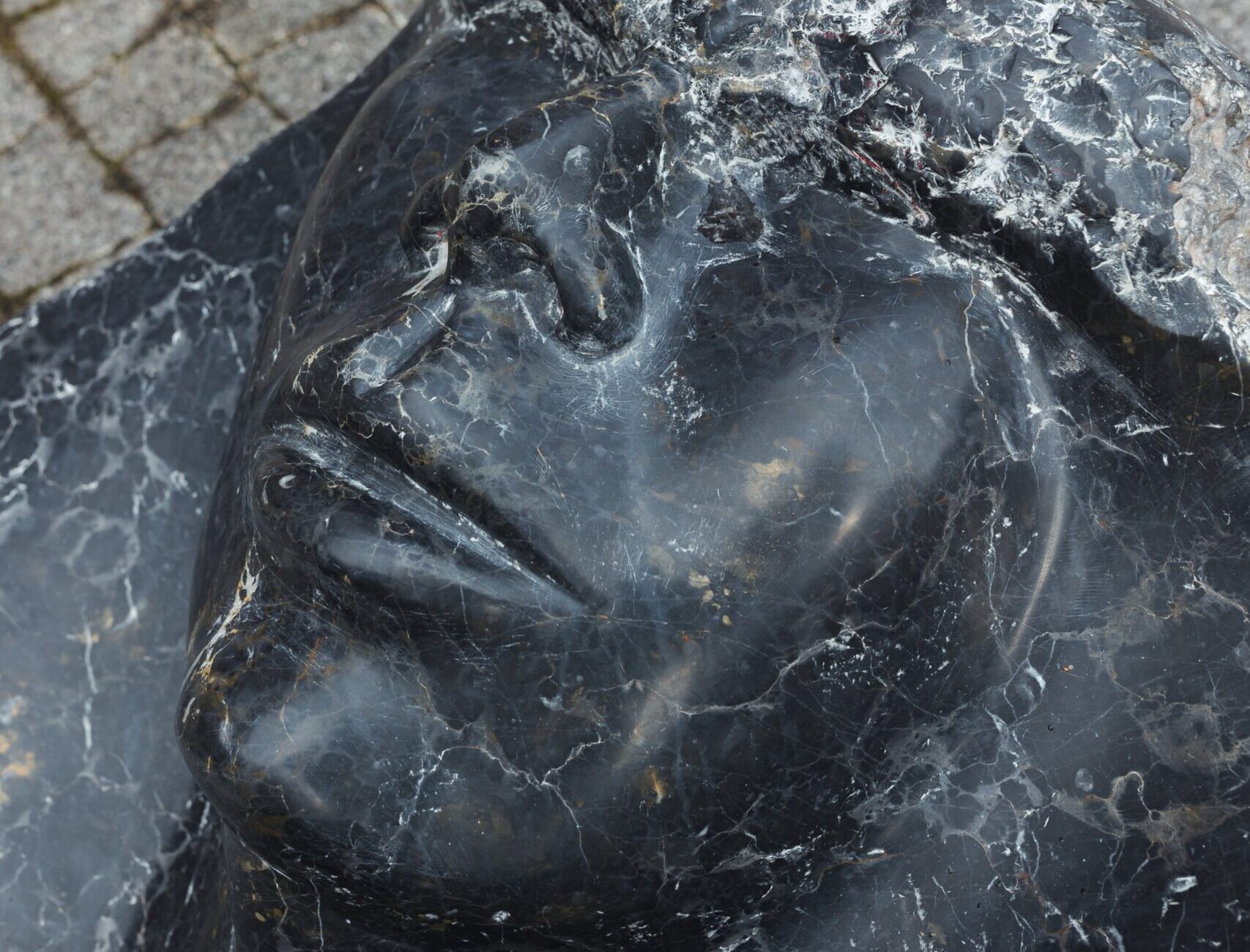 Zbliżenie na czarne, białymi żyłkami żyłkowane marmurowe oblicze rzeźby leżącej — widoczny profil nosa, zamknięte oko i usta oraz fragment kostki brukowej w tle.