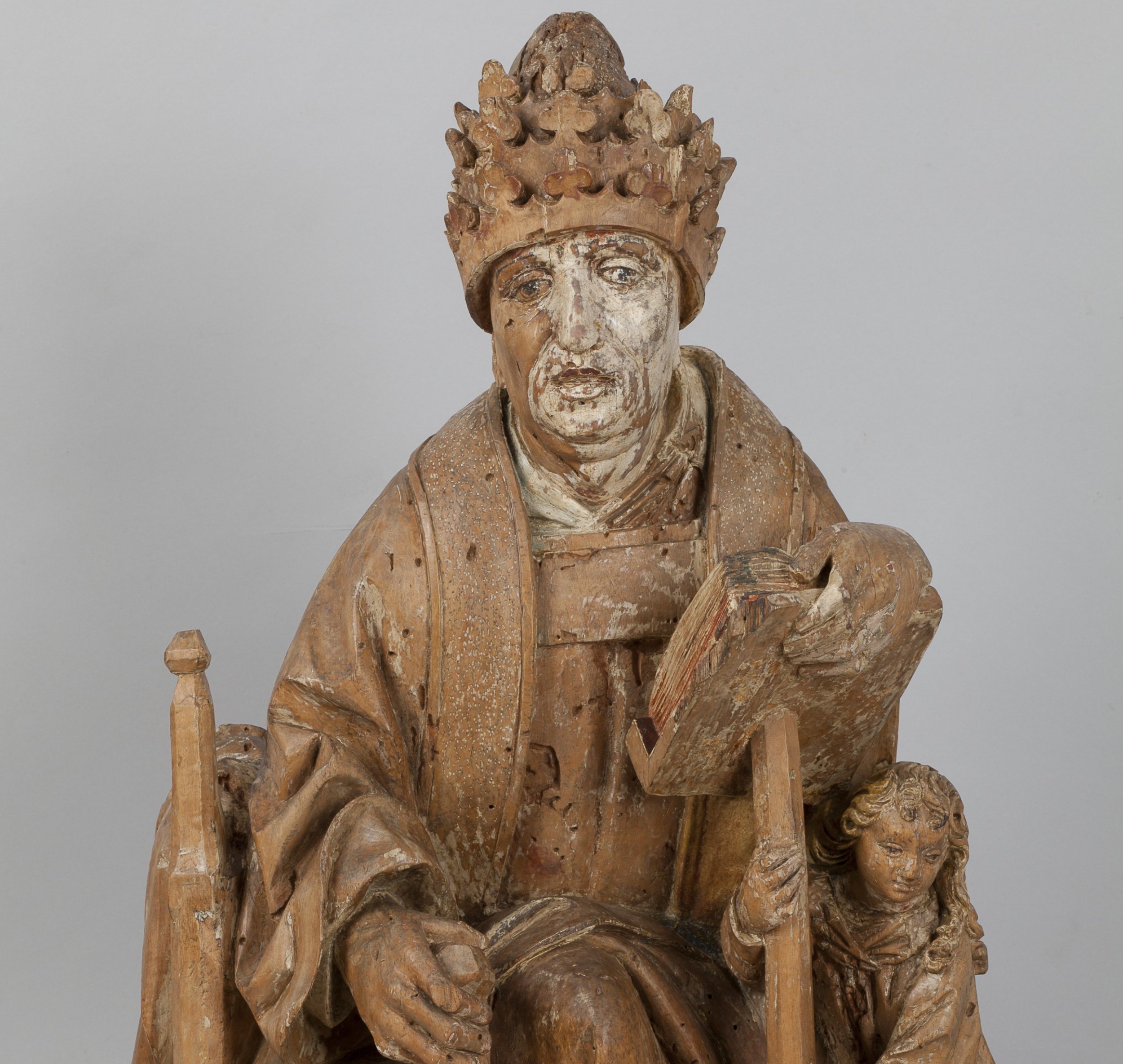Rzeźba z drewna przedstawiająca siedzącą, koronowaną postać w liturgicznych szatach, z jedną dłonią spoczywającą na otwartej książce opartej na pulpicie, wykonana w zużytym, miejscami spękanym polichromowanym drewnie, u podstawy po prawej mała figura anioła podpierającego pulpit.