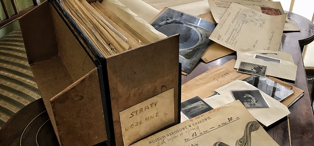 Na drewnianym stole kartonowe pudło archiwalne z widocznym napisem "STRATY WOJENNE" oraz rozrzucone po stole pożółkłe dokumenty, koperty, małe czarno-białe fotografie i formularz z nagłówkiem "MUZEUM NARODOWE W KRAKOWIE", w tle fragment pasiastego fotela.