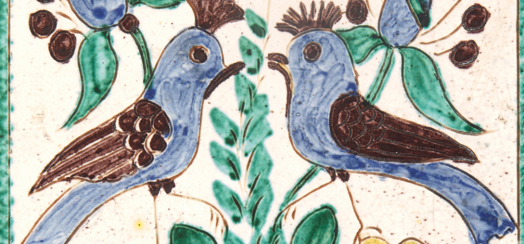 Glazurowany ceramiczny kafel przedstawiający dwa stylizowane niebieskie ptaki zwrócone twarzami do siebie, siedzące na gałązkach z zielonymi liśćmi i brązowymi owocami, z brązowymi piórami i grzebieniami oraz centralnym pionowym ornamentem roślinnym na jasnym tle.