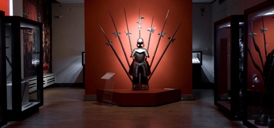 Wnętrze muzeum z centralnie ustawioną metalową zbroją na cokole przed czerwonym panelem, za którą wachlarzowo rozstawione długie bronie drzewcowe; po bokach przeszklone gabloty, drewniana podłoga i przytłumione oświetlenie.