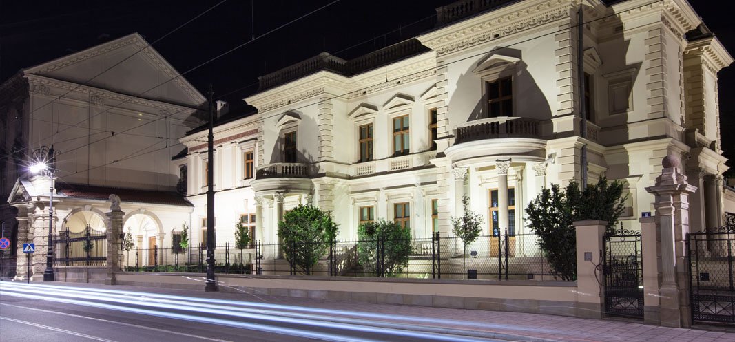 Oświetlony nocą jasny budynek o klasycystycznej elewacji z kolumnami, półokrągłymi balkonami i wieloma oknami, otoczony niskim ogrodzeniem i krzewami; po lewej arkadowe przejście i brama, a na pierwszym planie chodnik i ulica z poziomymi smugami świetlnymi przejeżdżających pojazdów, latarniami i wiszącymi przewodami trakcyjnymi na tle ciemnego nieba.