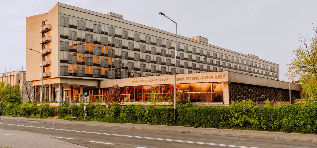 Modernistyczny wielopiętrowy budynek z równymi rzędami okien i przeszklonym parterem wspartym na kolumnach, z pomarańczowymi odbiciami w szybach, czerwonym graffiti na witrynach, zielonym żywopłotem i pustą ulicą z latarniami na pierwszym planie.