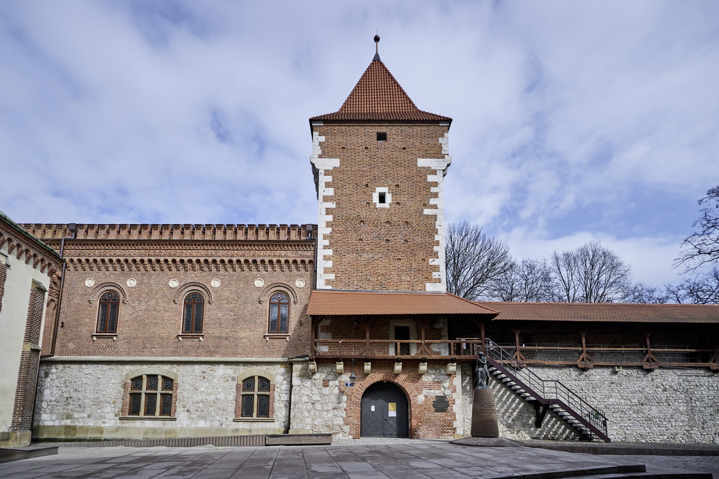 Centralna ceglana wieża z czerwonym dachówkowym dachem i białymi narożnikami, przylegająca do poziomego budynku z ozdobnym fryzem i trzema półokrągłymi oknami nad kamiennym cokołem, z drewnianym podcieniem i zewnętrznymi schodami po prawej oraz figurą na postumencie, a na pierwszym planie pusta przestrzeń wyłożona płytami i niebieskie niebo z chmurami oraz nagie drzewa w tle.