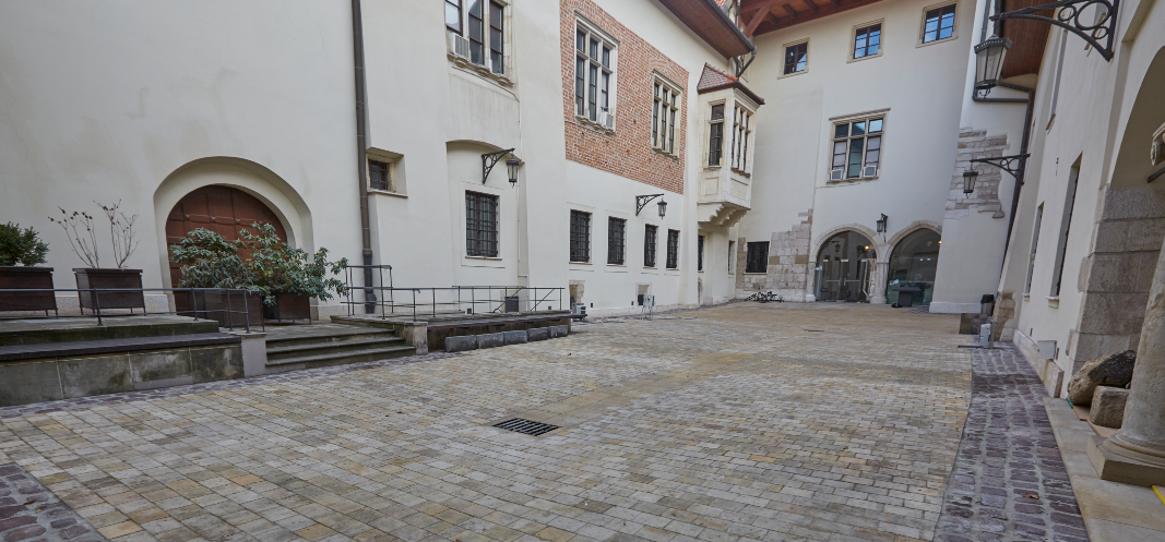 Pusty dziedziniec wyłożony jasnymi, kwadratowymi kamiennymi płytami, otoczony kremowymi i ceglanymi elewacjami z wieloma oknami, łukowymi przejściami i ozdobnym wykuszem, z kamiennymi schodami i donicami po lewej, metalowymi barierkami, lampami ściennymi i kratką odpływową pośrodku, na końcu przeszklone wejście i kilka zaparkowanych rowerów.