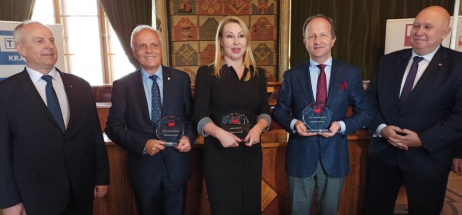 Pięć osób stojących w rzędzie w formalnych strojach w reprezentacyjnym, drewnianym wnętrzu; trzy środkowe osoby trzymają przezroczyste, okrągłe statuetki, w tle okna z zasłonami i ozdobna tapiseria.