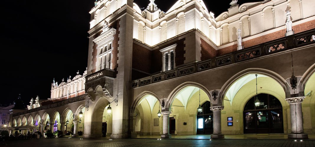 Oświetlona nocą frontowa fasada dużego, ozdobnego budynku z szeregiem kamiennych łuków i kolumn, wiszącymi lampami pod arkadami, dekoracyjnym gzymsem z rzeźbami na dachu i pustym brukowanym placem na pierwszym planie.