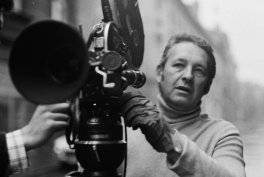 Czarno-białe zdjęcie osoby w golfie stojącej przy dużej, klasycznej kamerze filmowej na statywie na tle rozmytych budynków ulicy, z jedną ręką na kamerze i widoczną dłonią innej osoby po lewej.