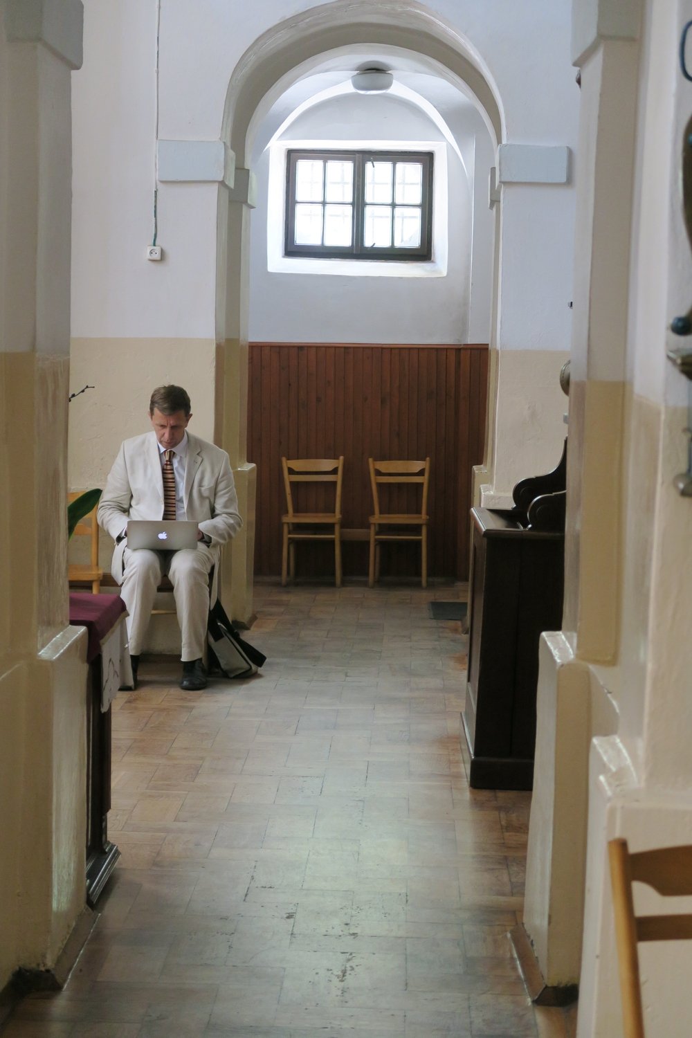 Wąski korytarz o białych ścianach i łukowym sklepieniu z małym prostokątnym oknem, drewnianą boazerią i dwiema pustymi krzesłami, w którym mężczyzna w jasnym garniturze siedzi na krześle z laptopem na kolanach i torbą obok, a na pierwszym planie widoczna długa drewniana podłoga.