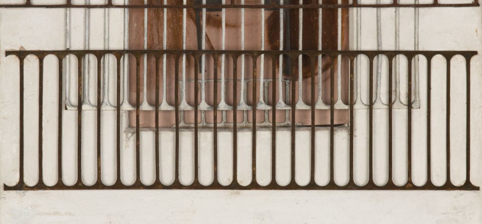 Fragment białej otynkowanej ściany z metalową, ciemnobrązową barierką balkonową ułożoną w dwóch poziomach z wąskimi pionowymi, zaokrąglonymi prętami, z widocznymi przebarwieniami rdzy i częściowo ukrytym za nią brązowym oknem.