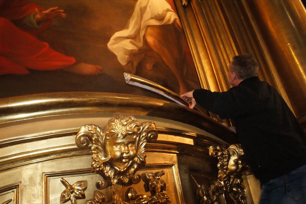 Mężczyzna w czarnej kurtce sięga narzędziem do odgiętego, złoconego gzymsu z rzeźbioną twarzą putta i roślinnymi ornamentami, pod fragmentem dużego malowidła i złotymi kolumnami.