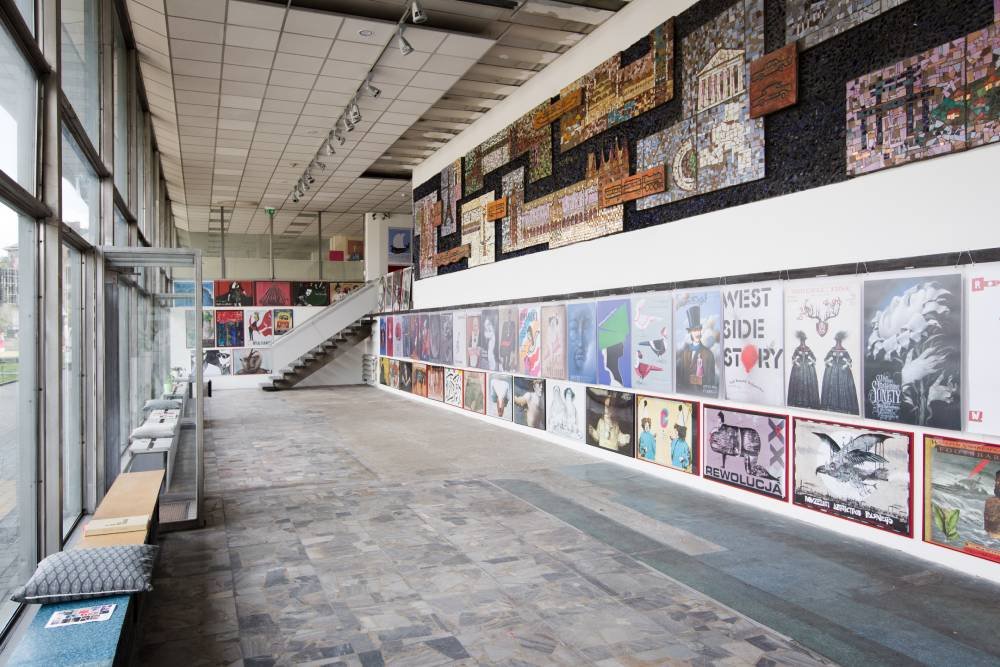 Wnętrze jasnej galerii z dużymi przeszklonymi ścianami po lewej, długą białą ścianą po prawej pokrytą rzędem plakatów i mozaikową instalacją nad nimi, schodami prowadzącymi na antresolę w tle, ławką z poduszkami przy oknach i kamienną posadzką.