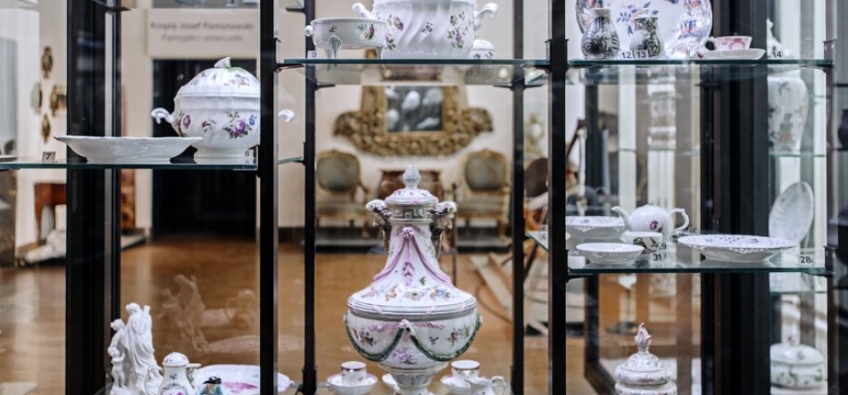 Witryna ekspozycyjna z czarną metalową ramą i szklanymi półkami wypełniona białą porcelaną z delikatnymi motywami kwiatowymi — centralnie ozdobny różowo-zielony wazon z pokrywką otoczony filiżankami, talerzykami, dzbankiem i figurkami, z rozmytym złoconym lustrem i klasycznymi fotelami widocznymi w tle.