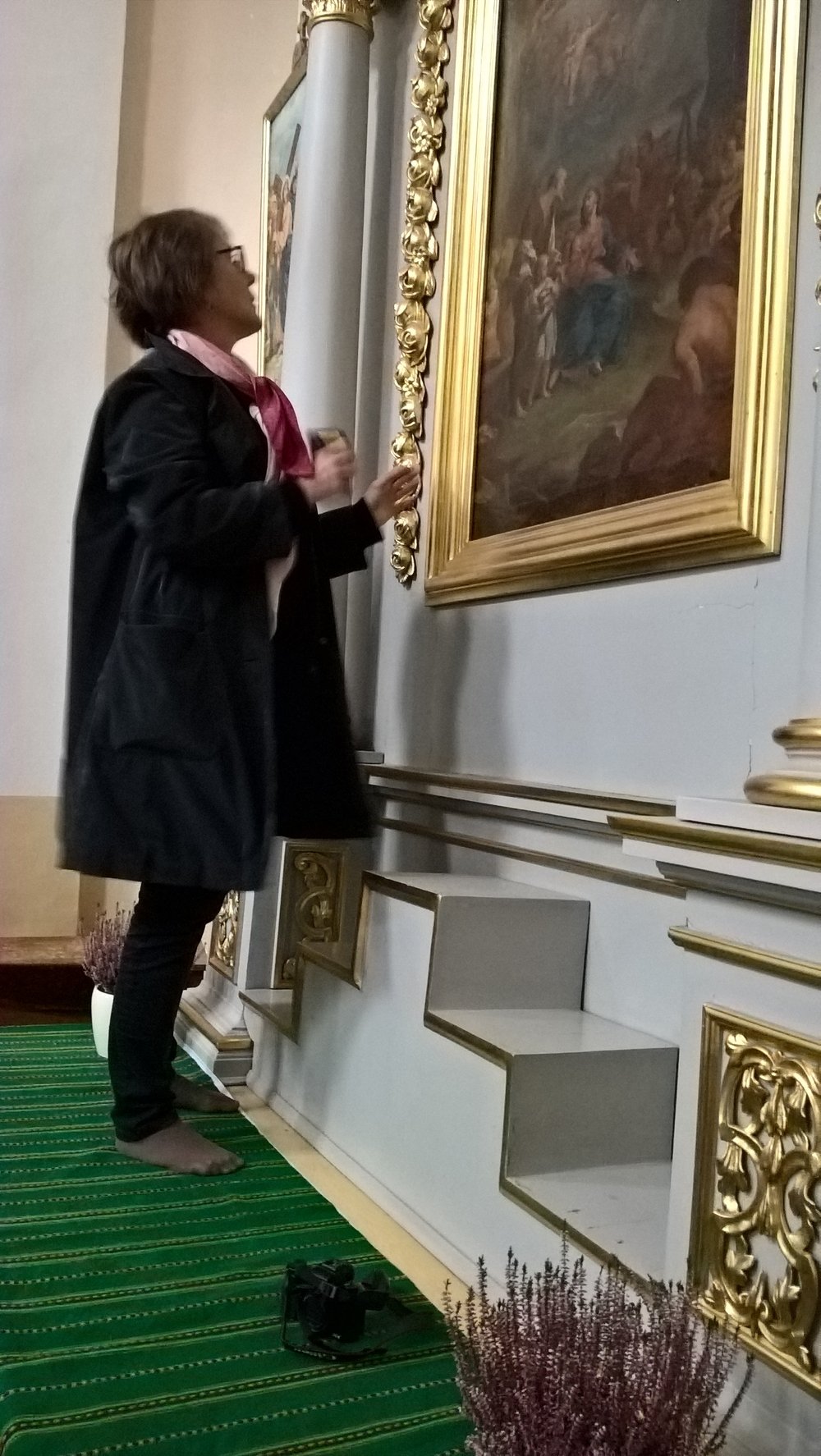 Osoba stojąca boso na zielonym, paskowanym dywanie, w ciemnym płaszczu i różowym szaliku, patrzy i dotyka bogato złoconej ramy dużego obrazu na ścianie; w otoczeniu widoczne zdobione kolumny, schodkowe podium, na podłodze leżą aparat fotograficzny i doniczka z wrzosem.