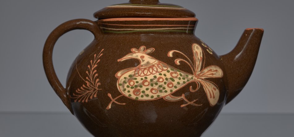 Błyszczący brązowy ceramiczny imbryk z pokrywką, uchwytem po lewej i dziobkiem po prawej, ozdobiony na środku korpusu kremowo‑pomarańczowo‑zielonym stylizowanym ptakiem z motywami roślinnymi oraz cienkimi paskami przy krawędzi pokrywki, na neutralnym szarym tle.