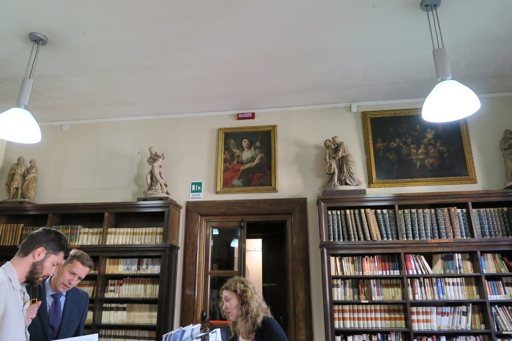 Wnętrze biblioteki z wysokimi drewnianymi regałami pełnymi książek, nad którymi wiszą oprawione obrazy i stoją rzeźby, przeszklonymi drzwiami pośrodku, zielonym znakiem wyjścia, dwiema wiszącymi białymi lampami sufitowymi oraz trzema osobami przy ladzie — dwoma mężczyznami po lewej patrzącymi na dokumenty i kobietą stojącą za ladą.