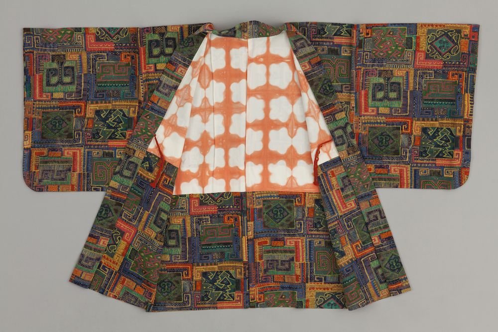 Otwarte, rozłożone na płasko kimono z szerokimi rękawami; zewnętrzna tkanina w wielobarwny geometryczny wzór kwadratów i labiryntów w zieleni, niebieskim, pomarańczowym i fioletowym, a wnętrze podszyte białą tkaniną z powtarzającym się pomarańczowym wzorem o zaokrąglonych, krzyżowych formach.