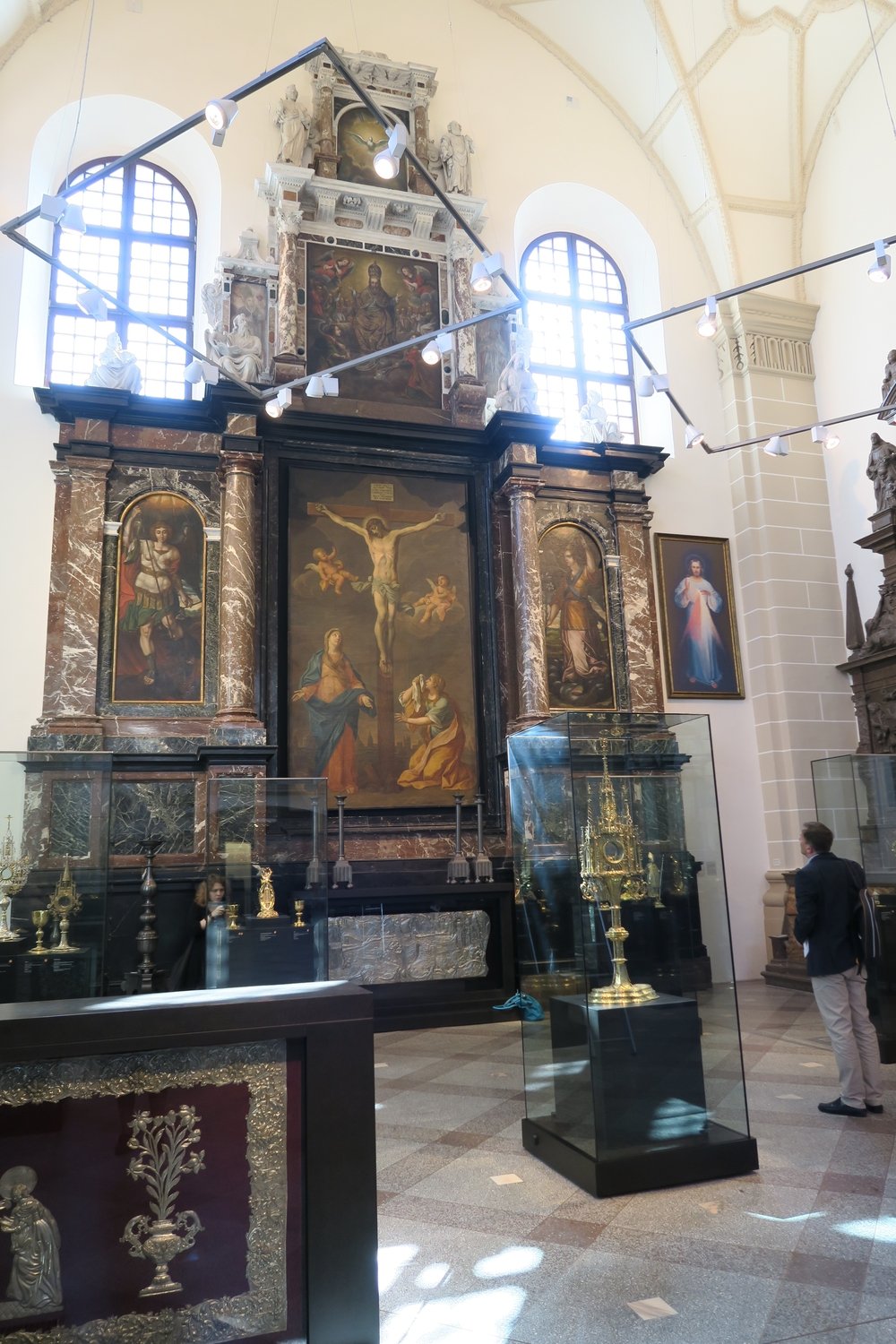 Wysokie, jasno oświetlone wnętrze kościoła z dużym marmurowym ołtarzem i centralnym obrazem Ukrzyżowania, flankowanym dwoma mniejszymi obrazami, kolumnami i rzeźbami u góry, dużymi oknami za ołtarzem oraz szklanymi gablotami z pozłacanymi relikwiarzami na pierwszym planie i osobą stojącą po prawej stronie.