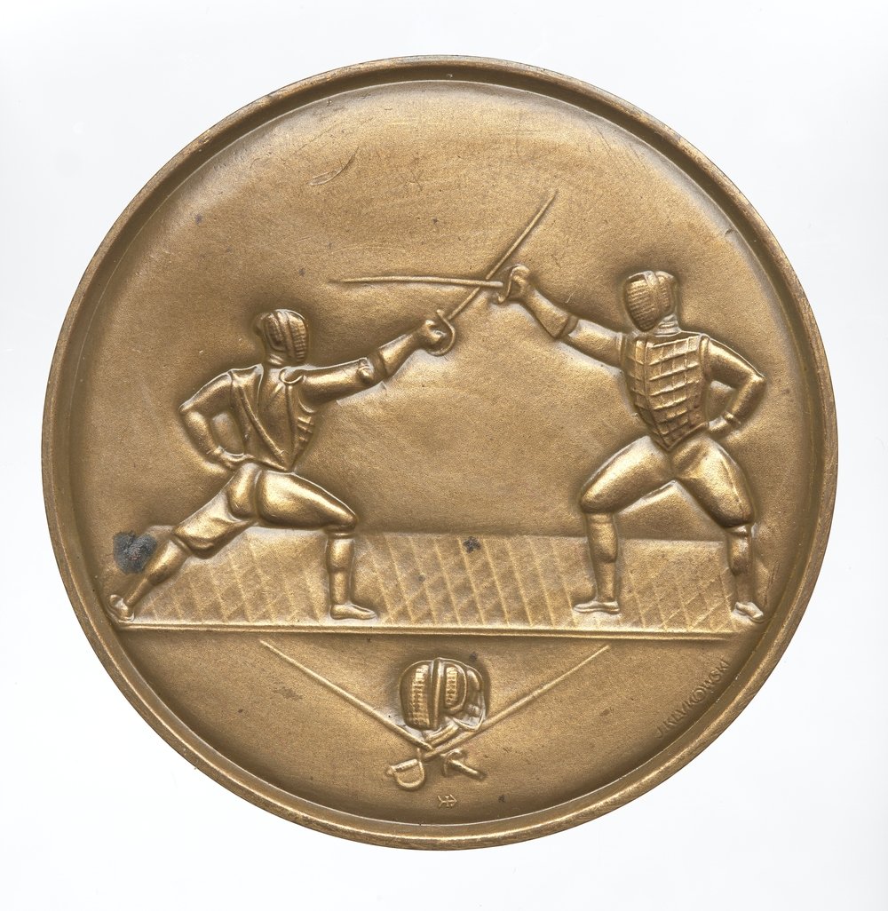 Okrągły brązowy medal z wytłoczonym reliefem dwóch zamaskowanych szermierzy w pozycji ataku stojących na poprzecznej planszy, ich szermiercze miecze skrzyżowane nad planszą, a w dolnej części mniejszy relief z skrzyżowaną bronią szermierską i maską.