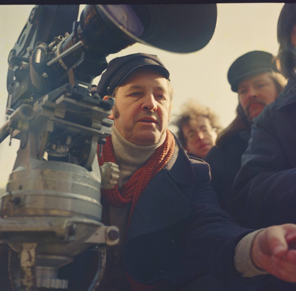 Mężczyzna w ciemnej czapce i płaszczu z beżowym golfem i rudawym szalikiem siedzi przy dużej kamerze filmowej na statywie, a za nim w tle widać dwóch innych mężczyzn w czapkach.