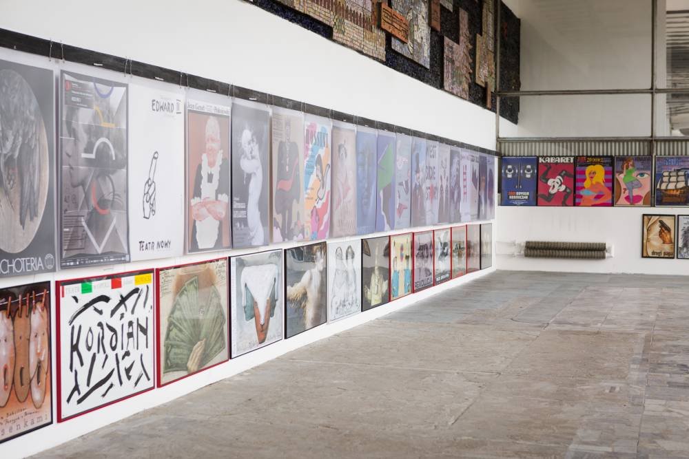 Wnętrze dużej, przemysłowej galerii z długą białą ścianą pokrytą dwiema rzędami różnokolorowych plakatów i grafik, betonową posadzką, wysokim sufitem z widocznymi belkami, mozaiką zawieszoną wysoko na ścianie i grzejnikiem w głębi.