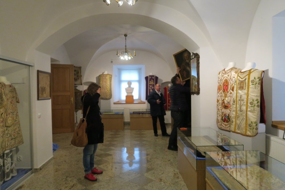 Jasne, sklepione pomieszczenie wystawowe z przeszklonymi gablotami i manekinami prezentującymi haftowane szaty oraz popiersiem na parapecie; trzy osoby w środku — kobieta z brązową torbą i czerwonymi butami patrzy, a dwóch mężczyzn dotyka dużego oprawionego portretu na ścianie.